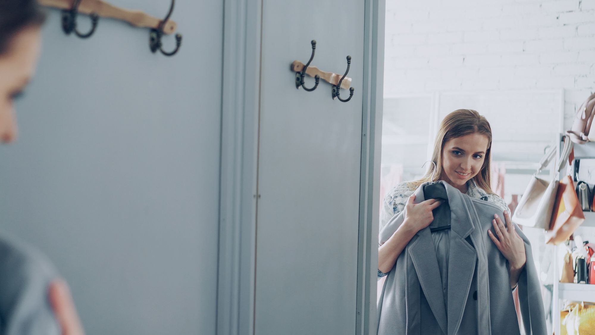 A person holding a grey coat, with handbags on a shelf